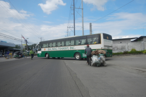 Ein grüner und weißer Bus fährt auf einer Straße neben einem Gebäude, mit Fußgängern und einem Radfahrer in der Nähe, Strommasten, Gebäuden, Bäumen und einer bewölkten Himmel.