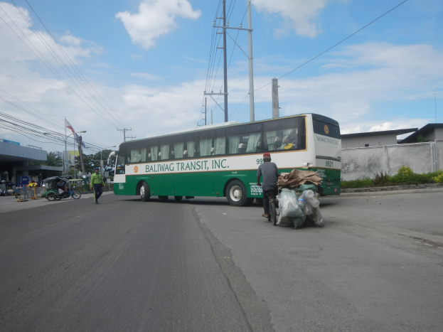 Ein grüner und weißer Bus fährt auf einer Straße neben einem Gebäude, mit Fußgängern und einem Radfahrer in der Nähe, Strommasten, Gebäuden, Bäumen und einer bewölkten Himmel.
