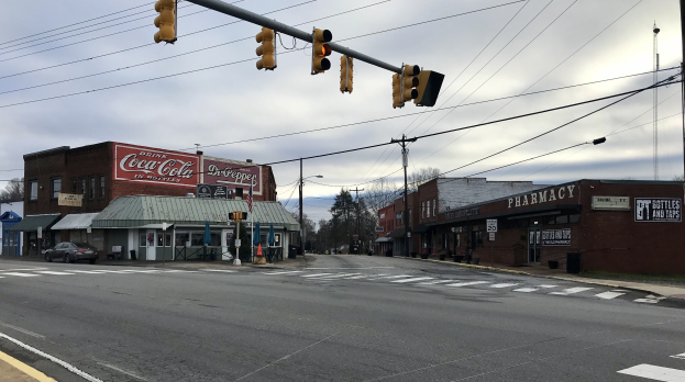 Stadtstraßenkreuzung mit einer Ampel, Fahrzeuge, Gebäude mit Schildern, Strommasten mit Kabeln, Bäume und ein bewölkter Himmel.