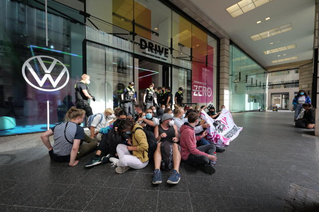 Menschen sitzen vor einem Volkswagen-Autohaus auf dem Boden und halten eine Fahne während einer Protestaktion.