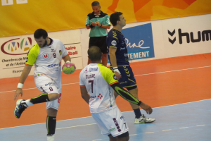 Eine Gruppe von Männern, die auf einem Handballfeld spielen, mit einem Mann, der einen Ball hält, und Schildern mit Text im Hintergrund.