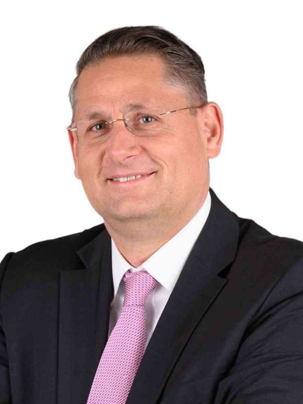 Ein lachender Mann im Anzug und Krawatte, der eine Brille trägt, vor einem weißen Hintergrund.