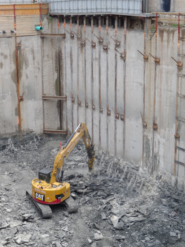 Bagger bei der Arbeit auf einer Baustelle mit Steinen und Eisenstangen neben einem teilweise gebauten Gebäude.