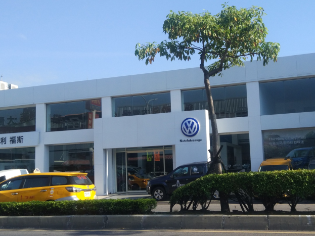 Volkswagen-Händler mit Glaswänden, parkenden Autos, umgeben von Pflanzen und einem Baum, unter einem sichtbaren Himmel.
