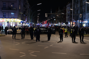 Eine Gruppe von Menschen geht nachts auf einer Straße in Berlin, Deutschland, während einer Demonstration, beleuchtet von Straßenlaternen und umgeben von Gebäuden, Bäumen, Lichtmasten, Verkehrszeichen und Schildern mit Text, mit Fahrzeugen auf der Straße und einem Banner mit Text im Hintergrund.