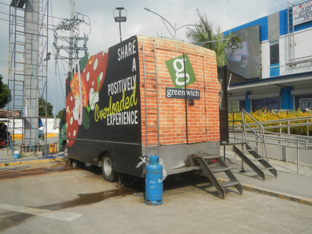 Ein weißer Lkw vor einem Gebäude mit einer Tafel, auf der "Teilen Sie eine positiv überladene Erfahrung" steht, mit Stufen, einem Zylinder auf dem Boden, Geländern, Strommasten mit Drähten, Laternen, Bäumen und einem bewölkten Himmel im Hintergrund.