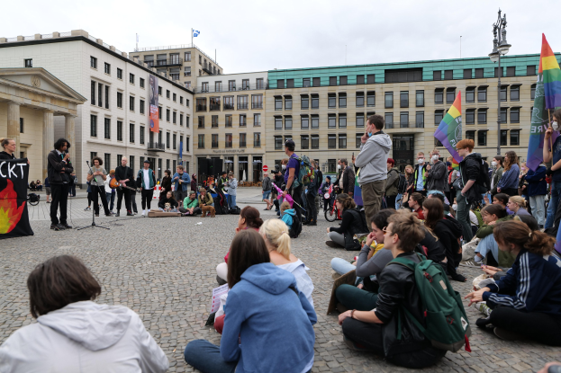 Eine Gruppe von Menschen, die auf dem Boden vor einer Menge sitzt, die Fahnen und Plakate hält, mit einer Person, die in ein Mikrofon spricht, einer Statue und Gebäuden im Hintergrund während einer anti-schwulen Demonstration in Berlin.