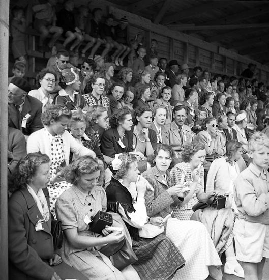 Schwarzes und weißes Foto einer Menge, die in einem Stadion ein Sportereignis ansieht, mit einer hölzernen Wand und einem Dach im Hintergrund.