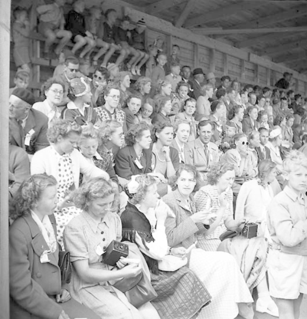 Schwarzes und weißes Foto einer Menge, die in einem Stadion ein Sportereignis ansieht, mit einer hölzernen Wand und einem Dach im Hintergrund.