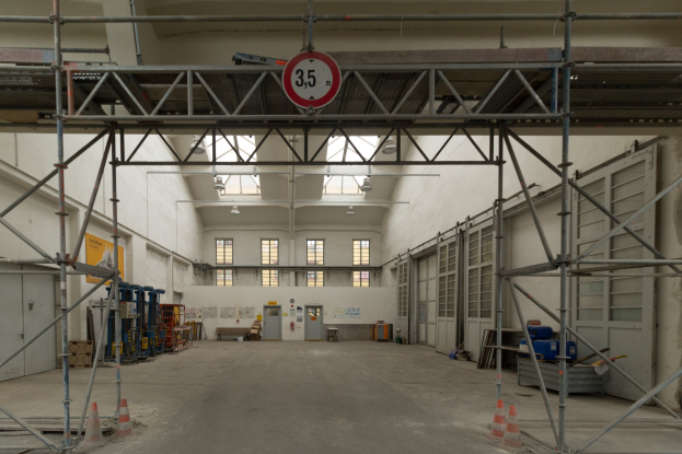 Lagerhalle mit ausgedehnten Gerüsten, Verkehrskegeln, Metallstangen, einem Schild, Maschinen, einer Tür, Fenstern, Wänden, Deckenleuchten und einem Dach.